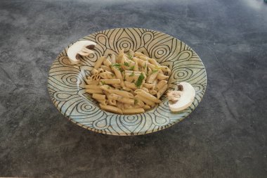 Üzerinde mantar olan bir kase makarna tezgahın üzerine konur. Makarna ortada dururken, mantarlar kasenin kenarına konur. Kase mavi ve beyaz bir tasarım.