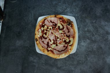 Lezzetli bir jambon ve ızgara pizza beyaz bir tabakta duruyor. Sos pizza boyunca cömertçe yayılıyor ve ağız sulandırıcı bir görünüm veriyor. Jambon eşit dağıtıldı.