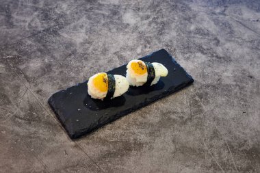 Siyah bir tabakta iki suşi rulosu. Ekmekler pirinç ve yosundan yapılmış. Tabak gri bir yüzeyde.