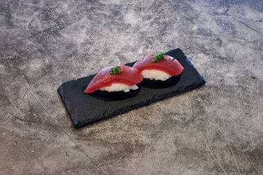 Altında siyah tabak olan bir tabak suşi. Suşi kırmızı ve beyaz. Suşinin üstünde yeşil bir garnitür var.