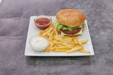 Beyaz tabakta patates kızartması ve ketçaplı bir hamburger. Patatesler kasede.