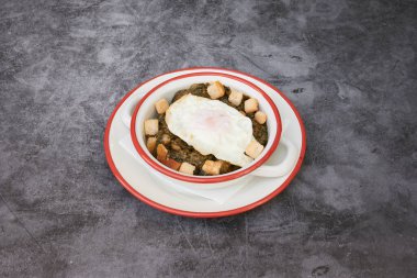 Kırmızı kenarlı beyaz bir tabak ve üzerinde de bir kase yiyecek. Yemek sebze ve yumurtanın karışımı.