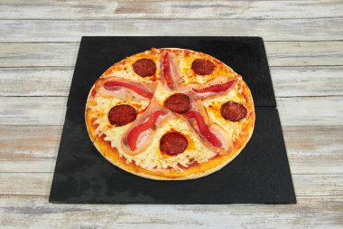 Sucuklu ve jambonlu bir pizza. Pizza siyah bir tabakta. Tabak ahşap bir masanın üzerinde.