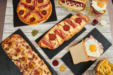 İçinde pizza, sandviç ve patates kızartması da dahil olmak üzere çeşitli yemek malzemelerinin olduğu bir masa. Ketçap ve hardal gibi baharatlar da var.