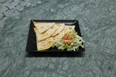 Üzerinde tortilla ve yanında salata olan bir tabak yemek. Tabak siyah, yemek sarı.