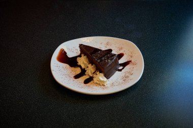 Üzerinde beyaz krema ve çikolata sosu olan lezzetli bir dilim çikolatalı pasta. Kek, siyah bir tezgahın üzerinde duran beyaz bir tabağa konur.