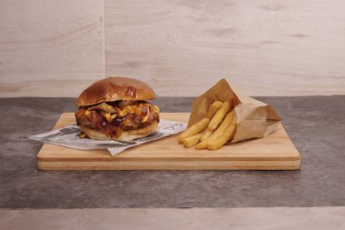 Peynirli ve pastırmalı bir hamburger ahşap bir kesme tahtasının üzerinde oturur. Hamburgerin yanında bir paket patates var.