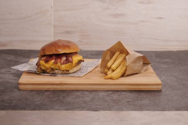Peynirli ve pastırmalı bir hamburger ahşap bir kesme tahtasının üzerinde oturur. Hamburgerin yanında bir paket patates var.