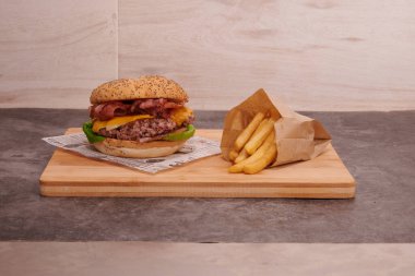 Peynirli ve pastırmalı bir hamburger ahşap bir kesme tahtasının üzerinde oturur. Hamburgerin yanında bir paket patates var.