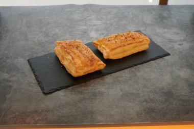 Siyah tabakta iki hamur işi. İkisi de kahverengidir ve tepesinde bir serpinti vardır. Tabak tezgahın üstünde.