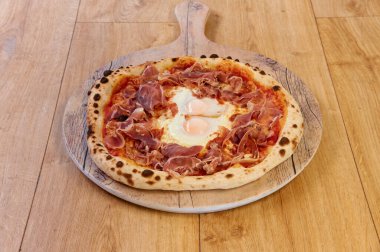 Lezzetli bir jambonlu ve yumurtalı pizza tahta bir tahtanın üzerinde duruyor. Pizza jambonlu ve yumurtalı ve çok iştah açıcı görünüyor. Tahta bu lezzetli yemek için mükemmel bir servis tepsisi.