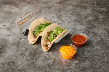 Üstünde yeşil marul ve soğan olan iki taco. Sarı ekşi krema kabı ve kırmızı salsa sosu.