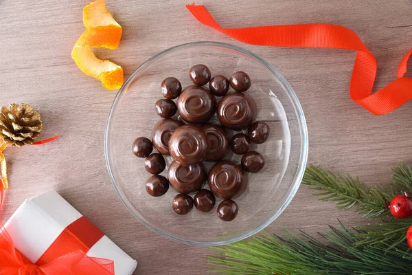 Chocolate candies in a glass transparent round dish in the center of a wooden table with Christmas decorations around. Top view.