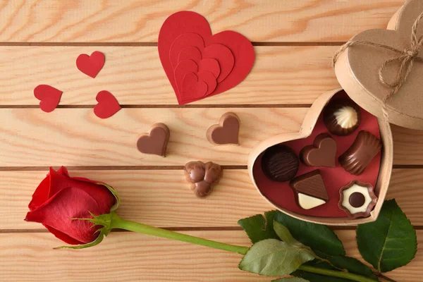 Box with an assortment of chocolates and red heart-shaped cutouts on a wooden slatted table and a red rose. Top view.
