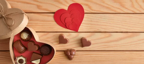 Box with assorted chocolates and heart-shaped red cut-outs on wooden slatted table. Top view.