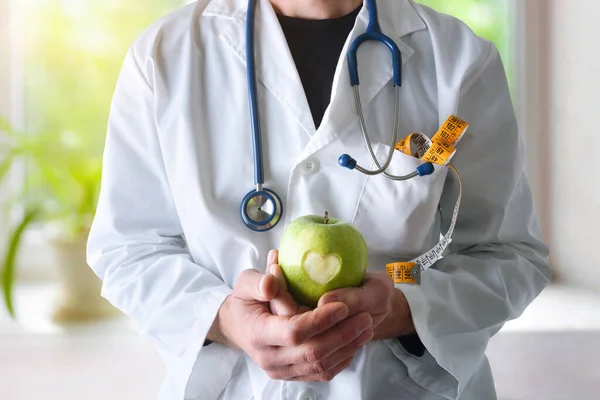 Dietician and nutritionist doctor with apple with a heart dressed in gown equipped with stethoscope and meter and window background with nature. Front view.