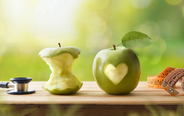 Healthy life and health check concept with eaten apple and whole apple with a bitten heart, stethoscope and tape measure on wooden table with nature background. Front view.