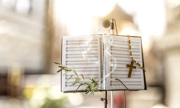Concept of choral songs for Palm Sunday celebration with music stand with score decorated with olive branch and religious cross and church background. Front view.