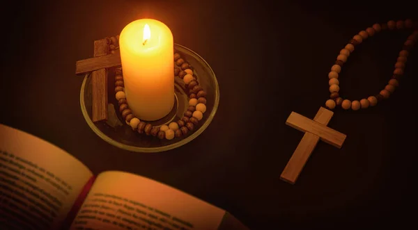 Christian spiritual environment for praying with candle illuminating the symbol of the cross and bible on black table at night. Top elevated view.