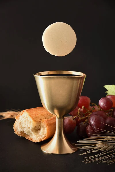 Reminder of the first communion with a chalice cup and a host on a black table with a loaf of bread bunch of grapes and ears of wheat. Vertical composition. Front view.