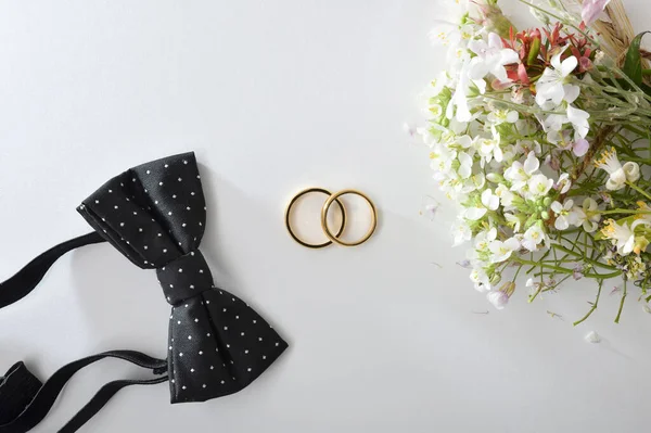 Wedding concept with gold rings in the center on white table and black bow tie and flower bouquet around. Top view.
