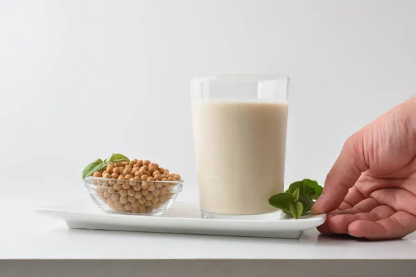 Detail of hand showing alternative soy drink on plate with full glass and bowl with dry beans on white table and white isolated background. Front view.