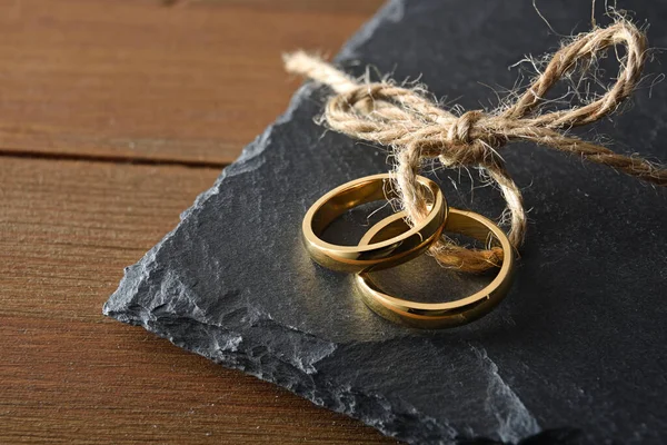 Detail of two gold rings joined with jute rope on slate stone on a rustic wooden table.. Top view.