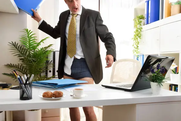 Businessman in online meeting in his home office surprised to be seen half dressed picking up a file cabinet.