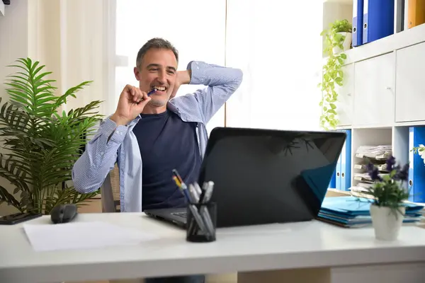 Freelance entrepreneur smiling biting a pen in his home office looking proudly at a laptop sitting on a chair in a white office with shelves full of folders and a window in the background.