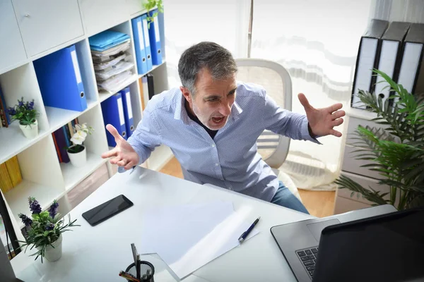 Angry man in front of a laptop sitting on a chair in a white office with shelves full of file cabinets elevated view
