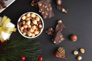 Fındıklı çikolata ve Noel 'de çam yaprağı ve çam kozalağı ile süslenmiş siyah bir masanın üzerinde kurumuş meyve. Üst görünüm.