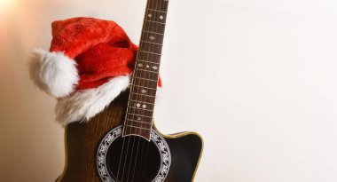 Akustik gitar gövdesi, izole edilmiş beyaz bir duvara yaslanmış Noel Baba şapkası. Noel gitarı müzik konsepti.