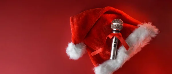 Christmas musical background with metallic stage microphone with red bow on santa hat isolated on red background. Top view.