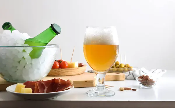 Fresh beer in a glass and container with ice full of bottles on white table with appetizer and white isolated background. Front view.