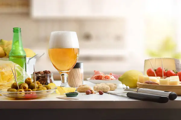 Appetizer with beer, olives, cheese and ham and cheese and other snacks on white kitchen bench and kitchen background behind. Front view.