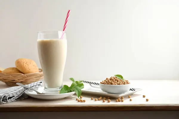 Glass of soy drink with red straw on white wooden table with bowl full of dry seeds and basket with bread with white isolated background. Front view.