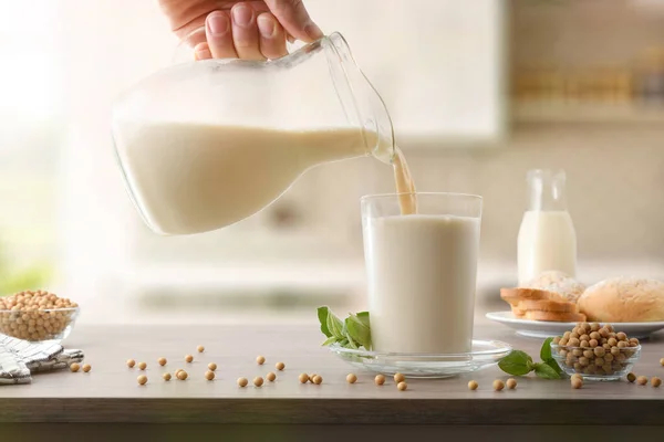 Filling glass with soy milk from a jug on wooden kitchen table with bowls full of seeds and plate with bread.  Front view.