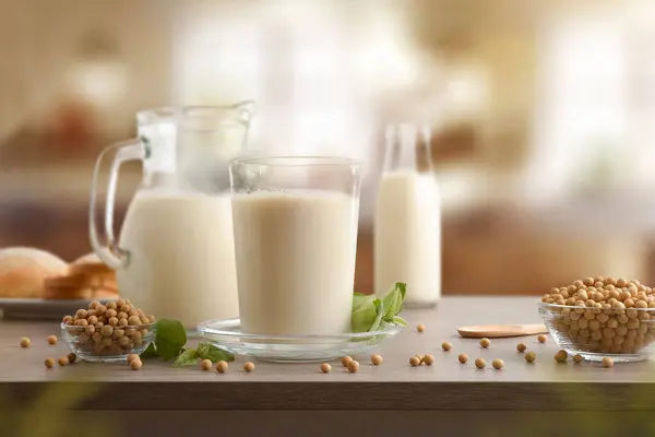 Glass containers with soy milk and grains in bowls on wooden table in a kitchen. Front view.