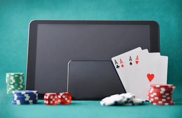 Electronic devices for online casino game on gaming table with chips, cards and dice on felt table and green background. Front view.