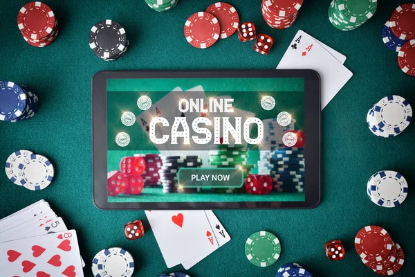 Concept of playing casino games online through an app with a tablet with a screen announcing that you play online casino on a table with green felt full of betting chips, cards and dice. Top view.