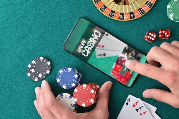 Online casino player entering app to play with hand pressing on smartphone screen on green felt gaming table with betting chips and casino game objects. Top view.