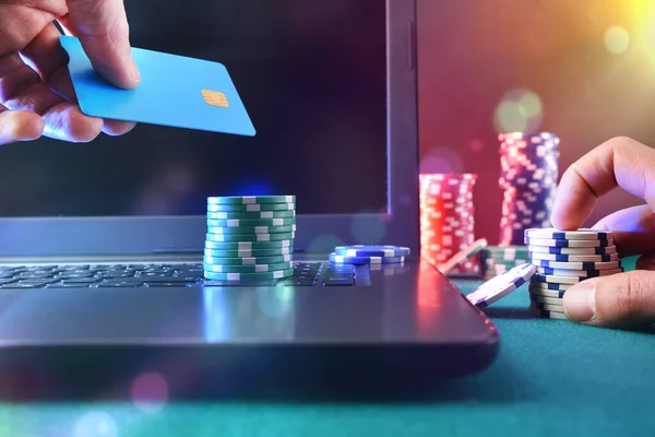 カジノゲームプレーヤーは ラップトップを使用して仮想ベットをするために支払いカードにアクセスします 片手のチップと もう片方の銀行カード フロントビュー ロイヤリティフリーのストック写真
