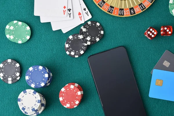Concept of playing casino games online with smartphone on a game table with green felt full of game objects and bank cards. Top view.