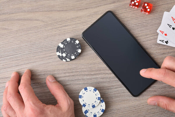 Online gambling concept with hand using smartphone on wooden table and game pieces. Top view.