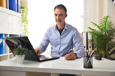 Mavi tişörtlü bir iş adamı bilgisayarda çalışıyor. Yazıyor ve ofiste notlar alıyor..