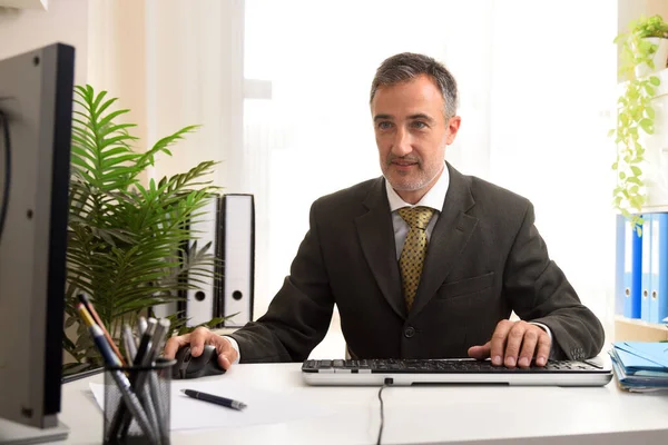 Businessman in gray suit and gold tie working with a computer typing and using the mouse in the office looking at a monitor.