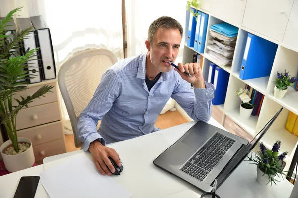 Pensative freelance entrepreneur biting a pen in his home office sitting on a chair in a white office with shelves full of folders and a window in the background. Elevated view.