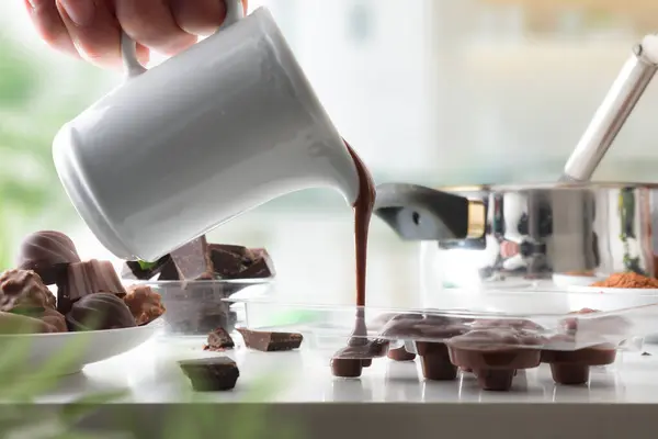 Person making chocolate bonbons on white kitchen bench at home with pouring hot chocolate into transparent mold. Front view.