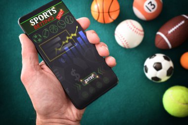 Farklı sporları temsil eden nesnelerin bulunduğu yeşil keçeli minderli bir masada akıllı telefondaki bir uygulamada online spor bilgilerine danışmanlık yapan kişi. Üst görünüm.