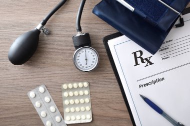 Üzerinde dokümanlar, tansiyon monitörü ve ahşap ofis masasında ilaçlar olan tansiyon reçeteli bir not. Üst görünüm.